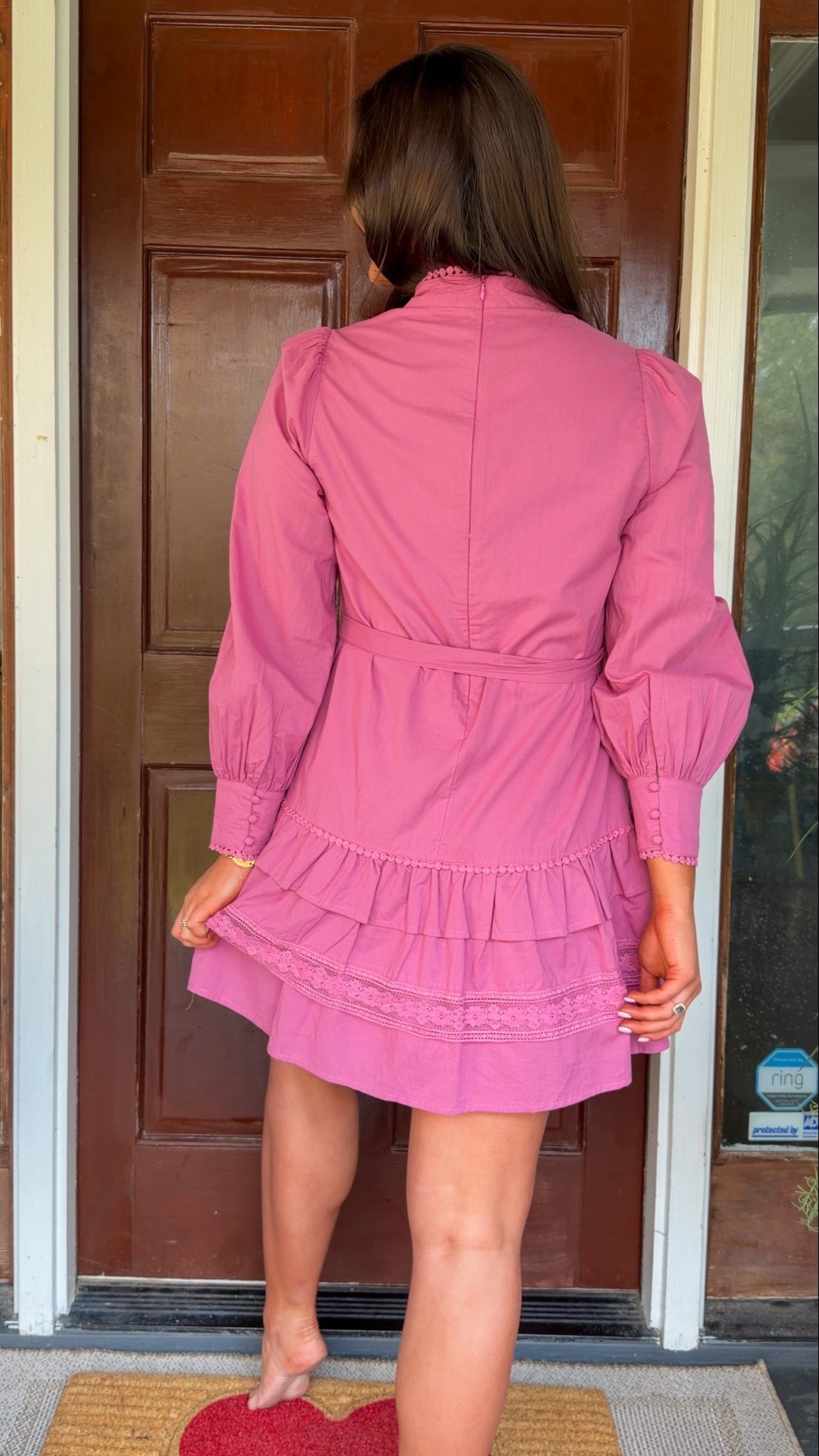 Mauve high-neck lace trim mini dress with long sleeves, self-tie waist, and tiered ruffle skirt worn by model standing in front of a wooden door