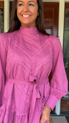 Mauve high-neck lace trim mini dress with long sleeves, self-tie waist, and tiered ruffle skirt worn by model standing in front of a wooden door