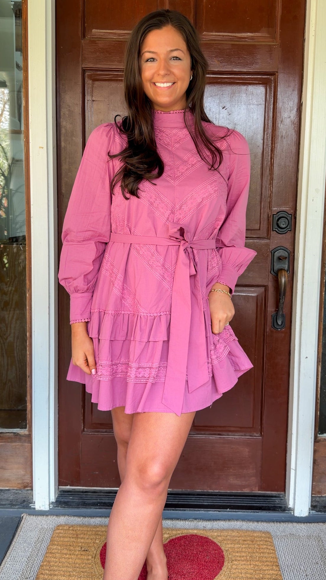 Mauve high-neck lace trim mini dress with long sleeves, self-tie waist, and tiered ruffle skirt worn by model standing in front of a wooden door