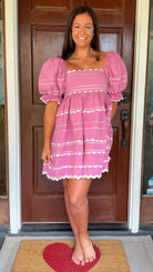 Model wearing a pink cotton poplin mini dress with white trim inset details, puff sleeves, and a square neckline, standing in front of a wooden door.