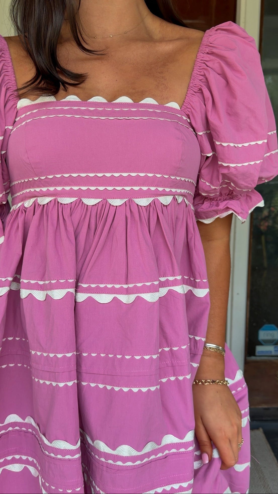 Model wearing a pink cotton poplin mini dress with white trim inset details, puff sleeves, and a square neckline, standing in front of a wooden door.