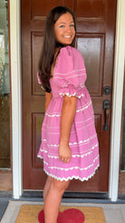 Model wearing a pink cotton poplin mini dress with white trim inset details, puff sleeves, and a square neckline, standing in front of a wooden door.