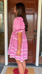 Model wearing a pink cotton poplin mini dress with white trim inset details, puff sleeves, and a square neckline, standing in front of a wooden door.