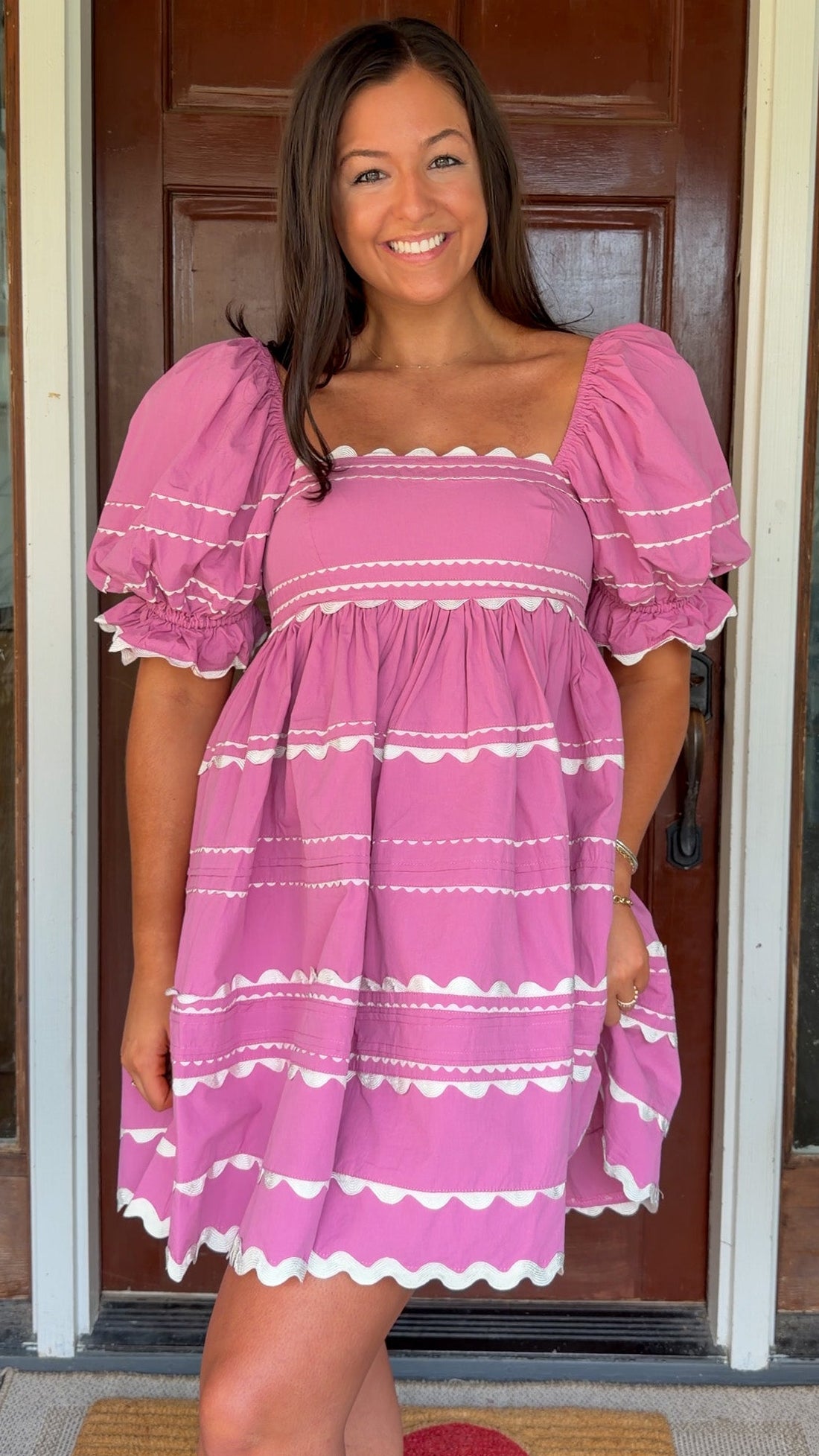 Model wearing a pink cotton poplin mini dress with white trim inset details, puff sleeves, and a square neckline, standing in front of a wooden door.