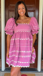 Model wearing a pink cotton poplin mini dress with white trim inset details, puff sleeves, and a square neckline, standing in front of a wooden door.