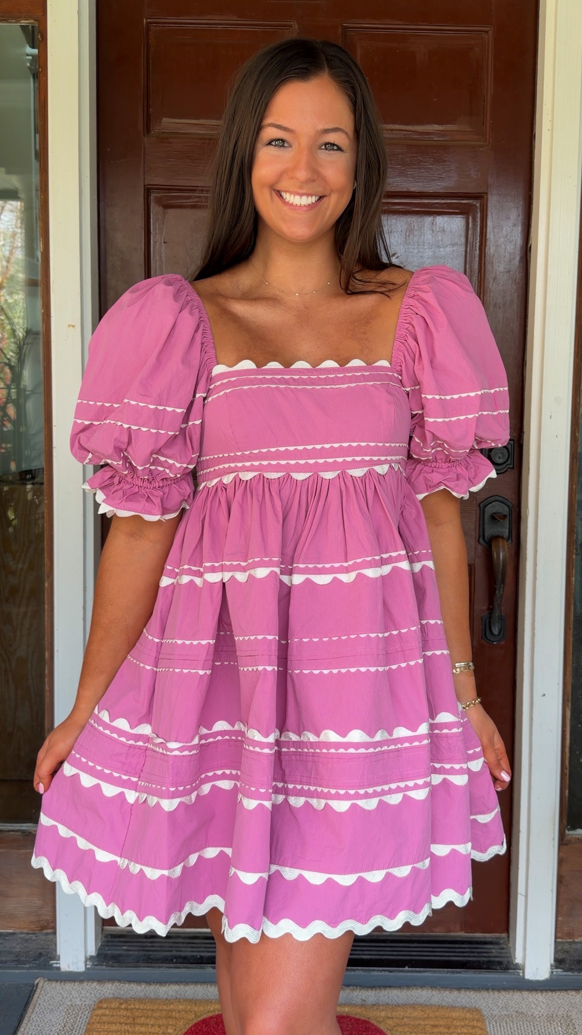 Model wearing a pink cotton poplin mini dress with white trim inset details, puff sleeves, and a square neckline, standing in front of a wooden door.