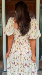 Model wearing a cream floral smocked midi dress with puff sleeves, flowy skirt, and side pockets, photographed in front of a wooden door.