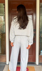 Model wearing a gray two-piece lounge set with lace trim cropped sweatshirt and matching elastic waist track pants featuring white side stripes and bow tie ankle details, standing in front of a wooden door.