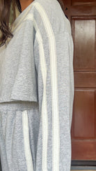 Model wearing a gray two-piece lounge set with lace trim cropped sweatshirt and matching elastic waist track pants featuring white side stripes and bow tie ankle details, standing in front of a wooden door.
