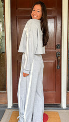 Model wearing a gray two-piece lounge set with lace trim cropped sweatshirt and matching elastic waist track pants featuring white side stripes and bow tie ankle details, standing in front of a wooden door.