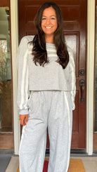 Model wearing a gray two-piece lounge set with lace trim cropped sweatshirt and matching elastic waist track pants featuring white side stripes and bow tie ankle details, standing in front of a wooden door.