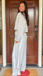 Model wearing a gray two-piece lounge set with lace trim cropped sweatshirt and matching elastic waist track pants featuring white side stripes and bow tie ankle details, standing in front of a wooden door.