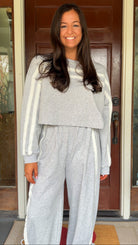 Model wearing a gray two-piece lounge set with lace trim cropped sweatshirt and matching elastic waist track pants featuring white side stripes and bow tie ankle details, standing in front of a wooden door.