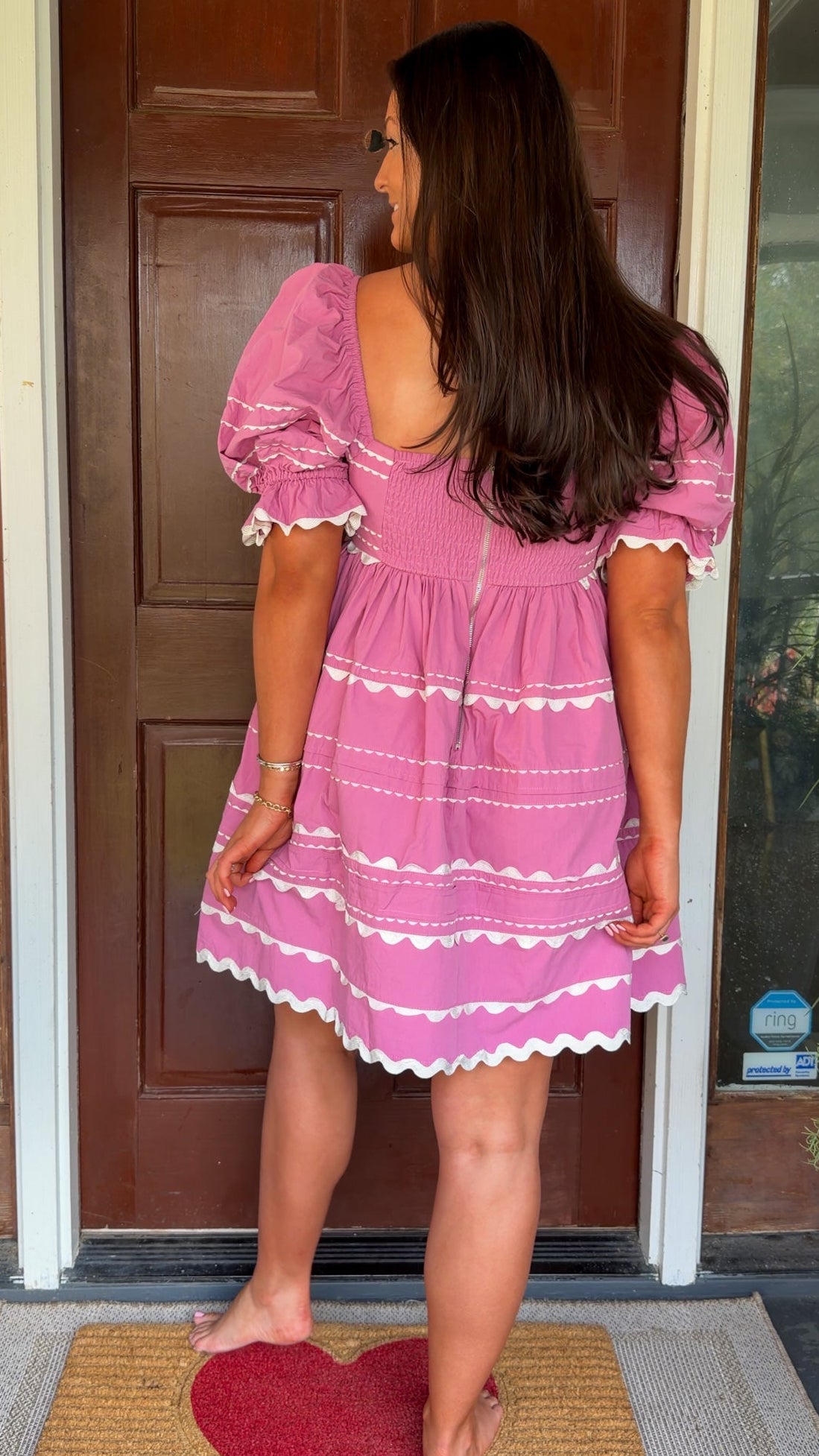 Model wearing a pink cotton poplin mini dress with white trim inset details, puff sleeves, and a square neckline, standing in front of a wooden door.