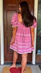 Model wearing a pink cotton poplin mini dress with white trim inset details, puff sleeves, and a square neckline, standing in front of a wooden door.