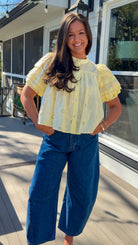 Woman wearing a yellow blouse and blue jeans standing on a wooden deck.