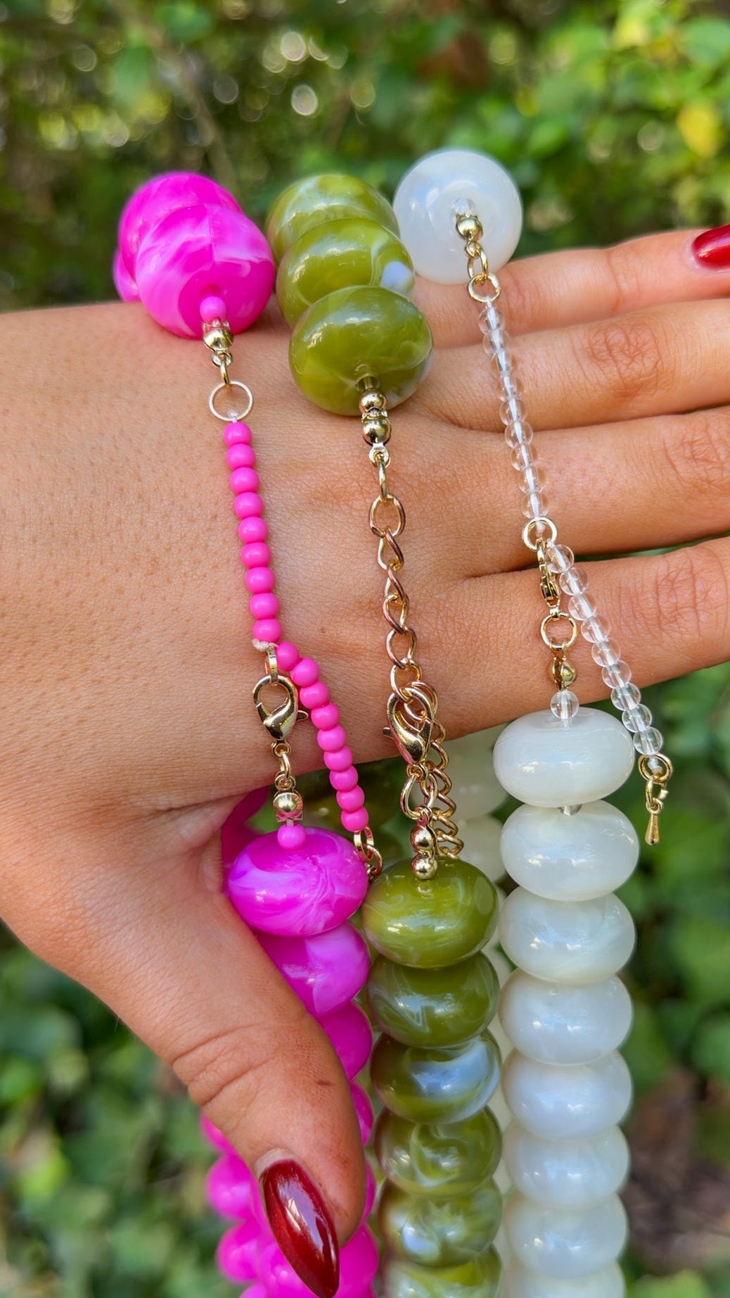 Three beaded prayer beads or necklaces held in a hand outdoors, featuring bright pink, olive green, and milky white beads with gold-toned chain details against a blurred green foliage background.