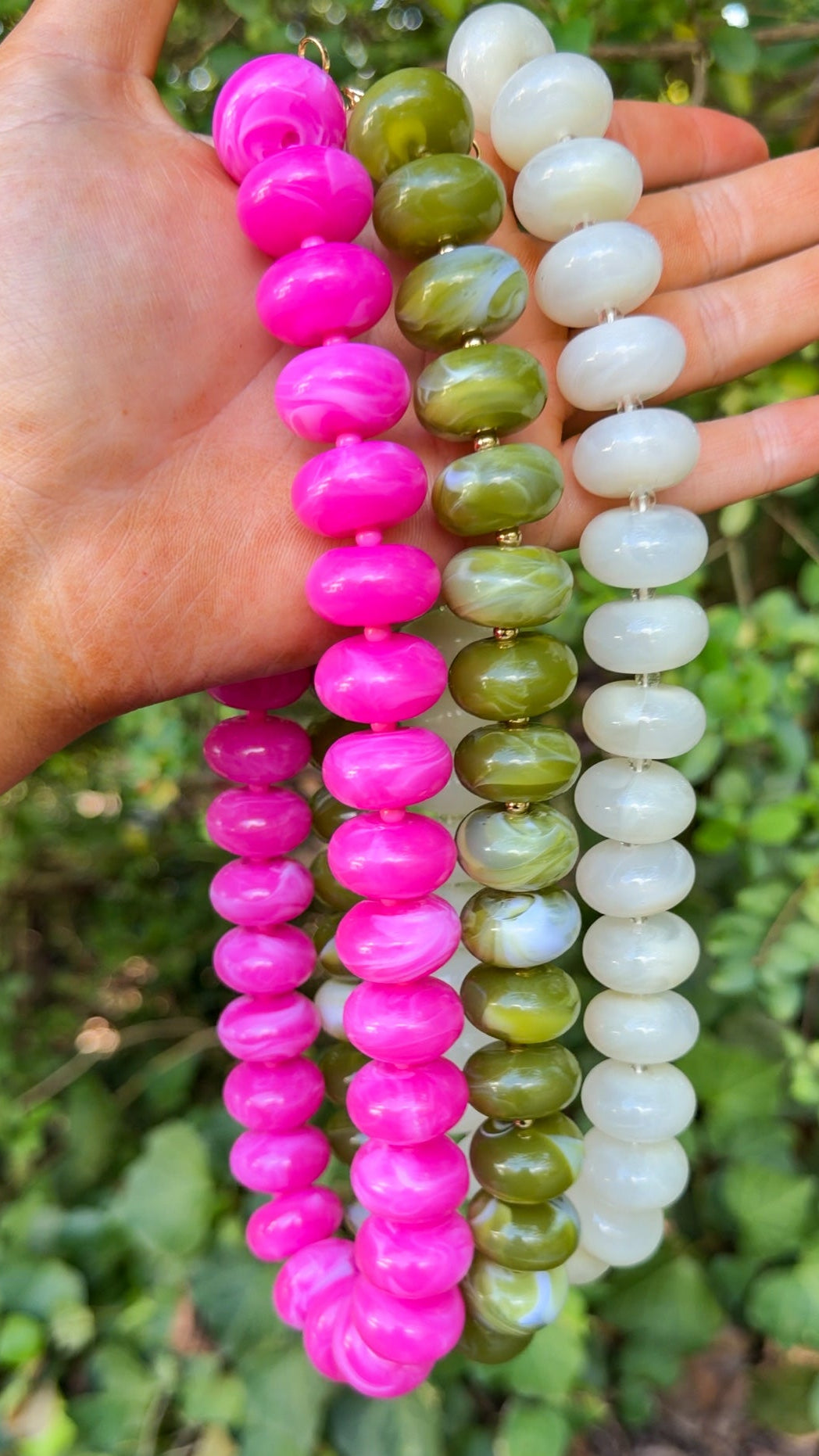 Three beaded necklaces held in a hand outdoors. One necklace is made of bright pink glossy round beads, one is made of olive green marbled round beads, and one is made of opaque white marbled round beads.