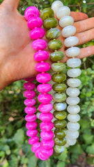 Three beaded necklaces held in a hand outdoors. One necklace is made of bright pink glossy round beads, one is made of olive green marbled round beads, and one is made of opaque white marbled round beads.