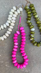 Three beaded necklaces laid flat on a gray concrete surface: one with large off-white oval beads, one with bright pink chunky oval beads and a short pink bead section with gold-tone hardware, and one with olive green marbled oval beads.