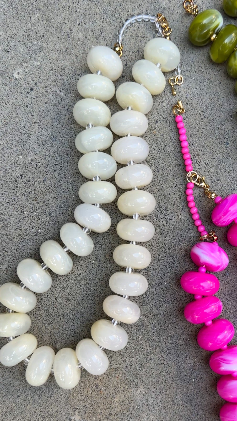 Two beaded necklaces laid flat on a gray concrete surface. One necklace features large, smooth, off-white disc-shaped beads with gold-toned chain links.