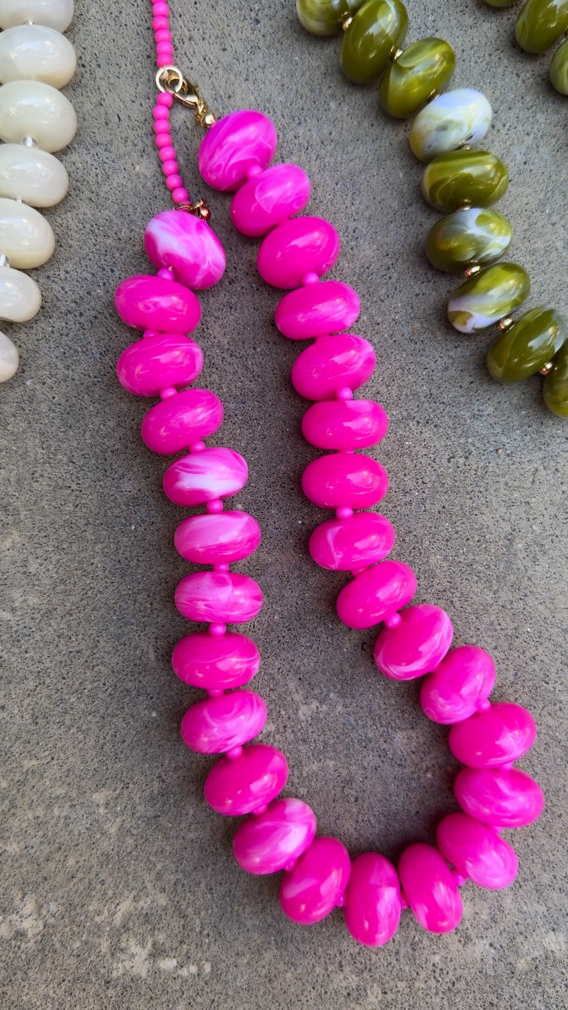A bright pink beaded necklace with chunky, rounded marbled beads, displayed on a gray textured surface alongside other colorful beads.