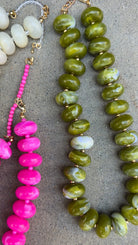Three beaded necklaces laid flat on a gray textured surface: one with large olive green marbled beads, one with bright pink beads and white beads, and a partial third necklace with cream beads at the top left.