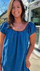 Blue denim mini dress with scoop neckline, short puff sleeves, and relaxed babydoll fit, worn outdoors in natural light.