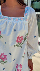 White embroidered mini dress with square neckline, long sleeves, and colorful floral embroidery, worn outdoors in natural light.