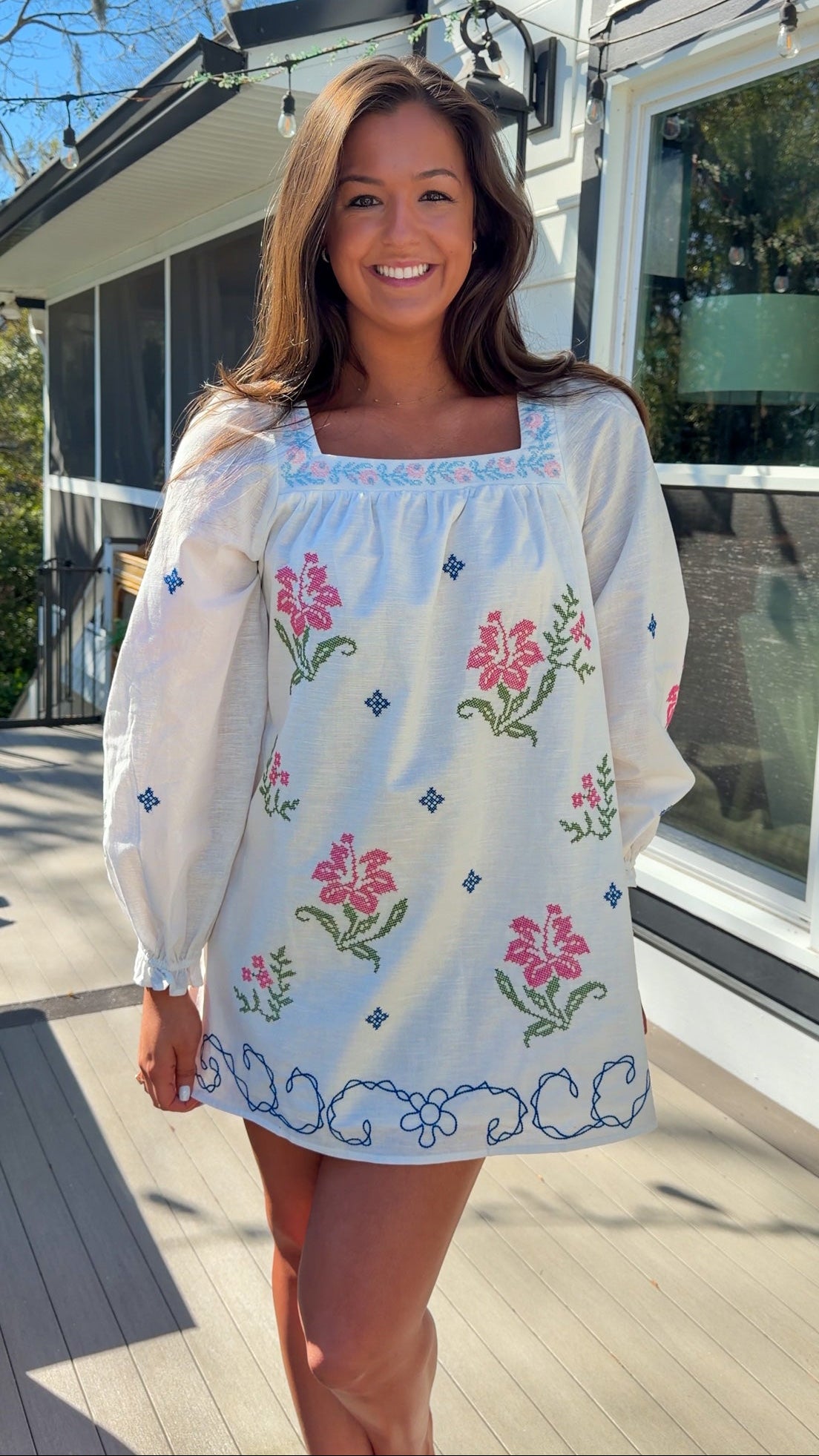 White embroidered mini dress with square neckline, long sleeves, and colorful floral embroidery, worn outdoors in natural light.