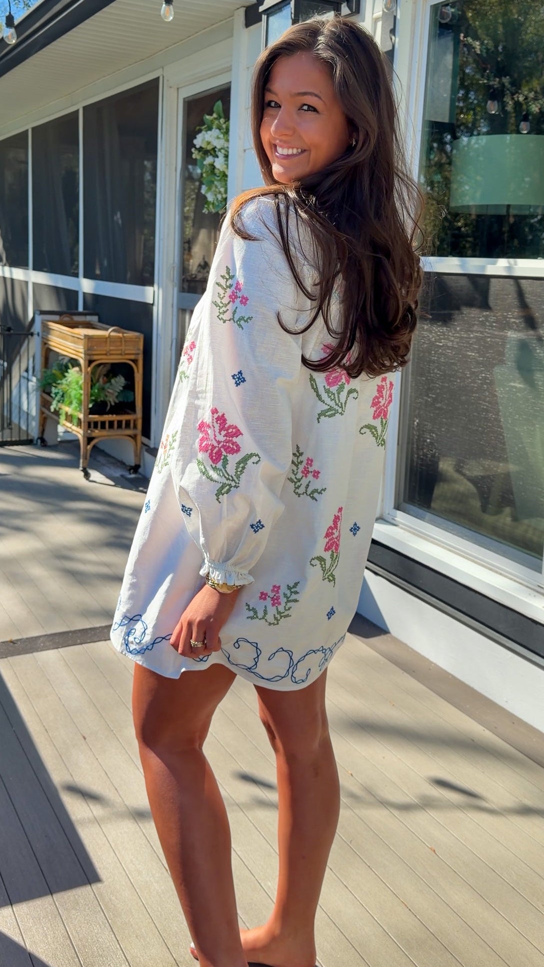 White embroidered mini dress with square neckline, long sleeves, and colorful floral embroidery, worn outdoors in natural light.