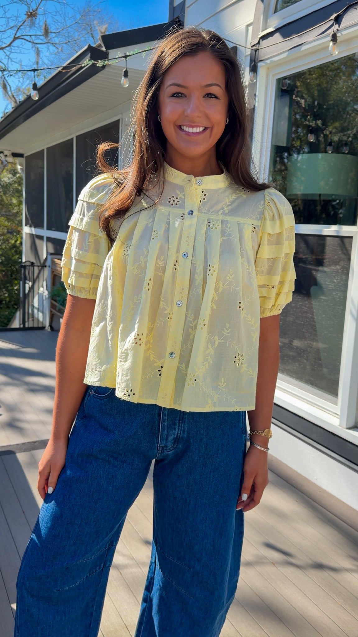 Woman wearing a yellow blouse with floral details and blue jeans standing outside a building.