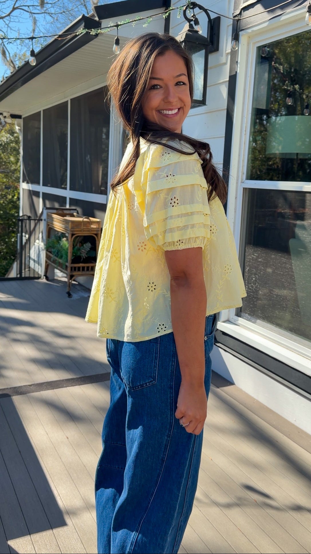 Woman wearing a yellow blouse and blue jeans standing on a wooden deck.