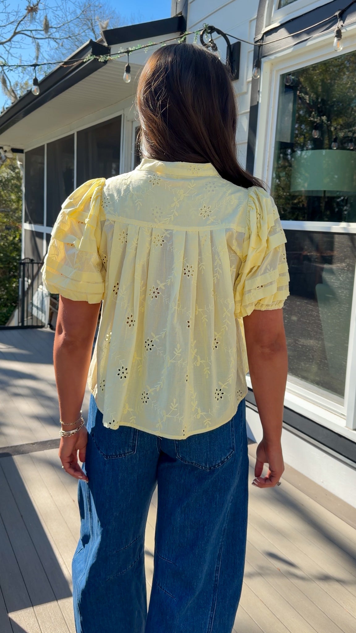 Person wearing a yellow blouse with floral patterns and blue jeans standing on a wooden deck.
