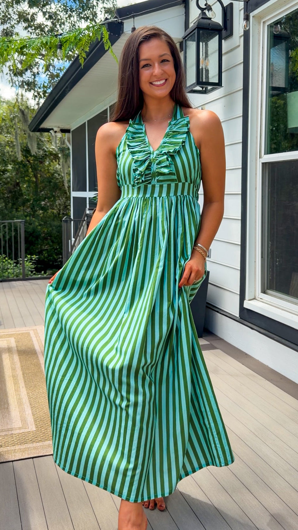 A woman wearing a green and white vertically striped halter-neck maxi dress stands on a wooden deck outside a house with white siding and black trim.