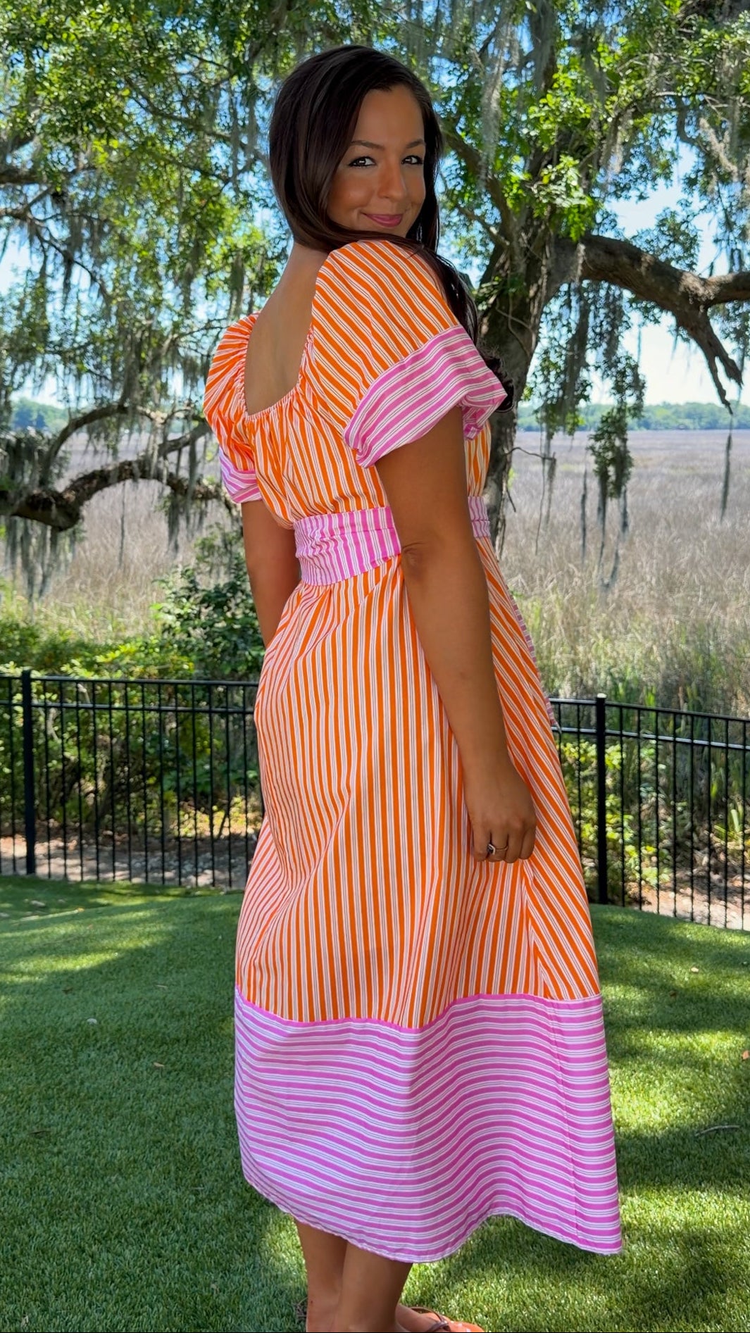 Woman wearing an orange and pink dress standing in a park with trees and grass.
