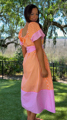 Woman wearing an orange and pink dress standing in a park with trees and grass.
