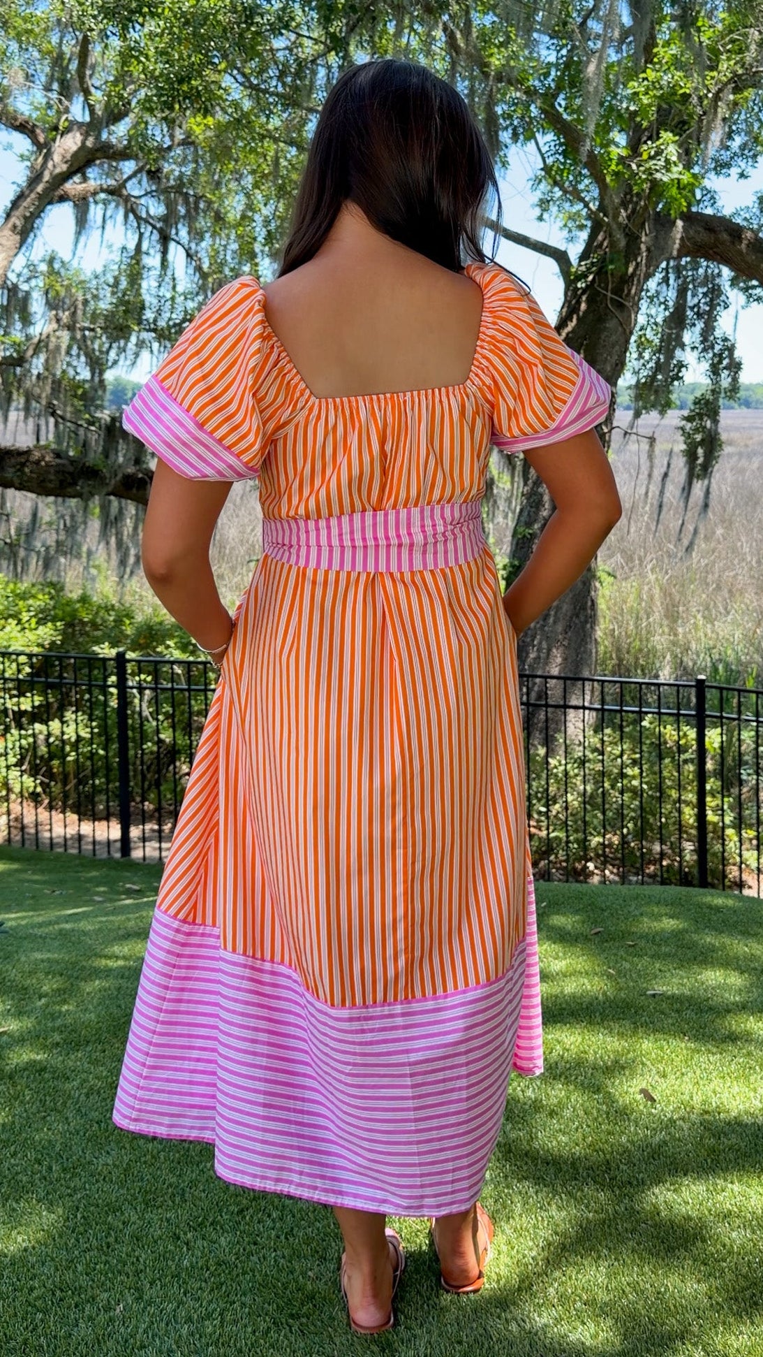Woman wearing a colorful dress with a striped pattern, standing outdoors with trees and grass in the background.