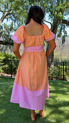 Woman wearing a colorful dress with a striped pattern, standing outdoors with trees and grass in the background.