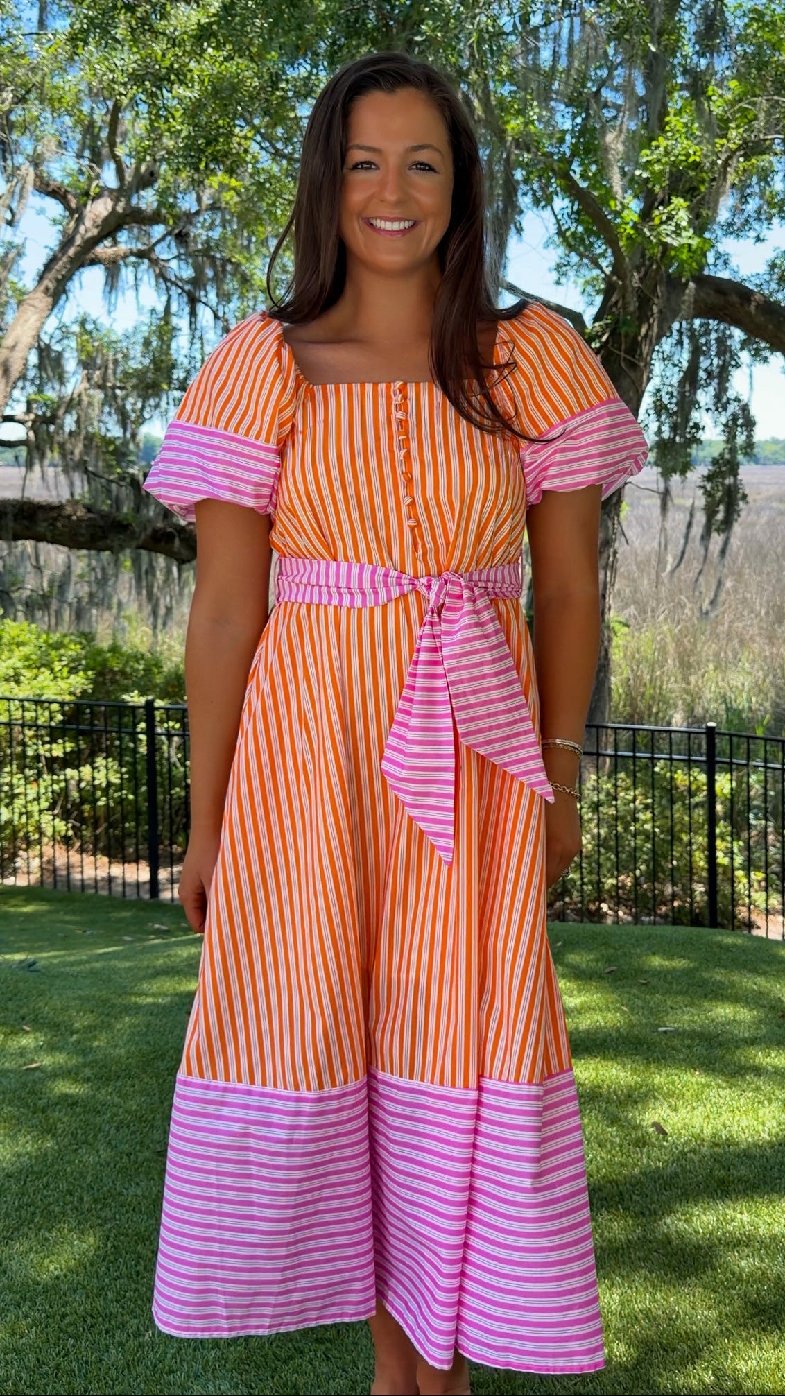 Woman wearing an orange and pink striped dress standing outdoors with trees and grass in the background
