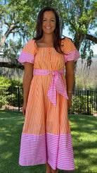 Woman wearing an orange and pink striped dress standing outdoors with trees and grass in the background