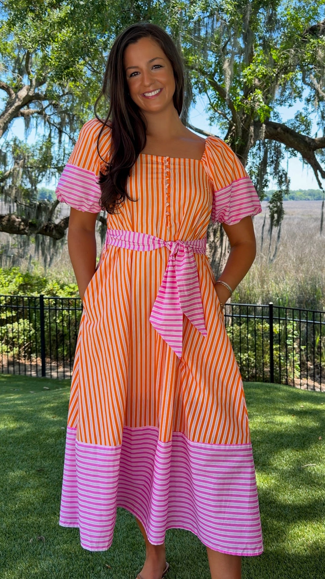 Woman wearing a colorful striped dress standing outdoors with trees and grass in the background