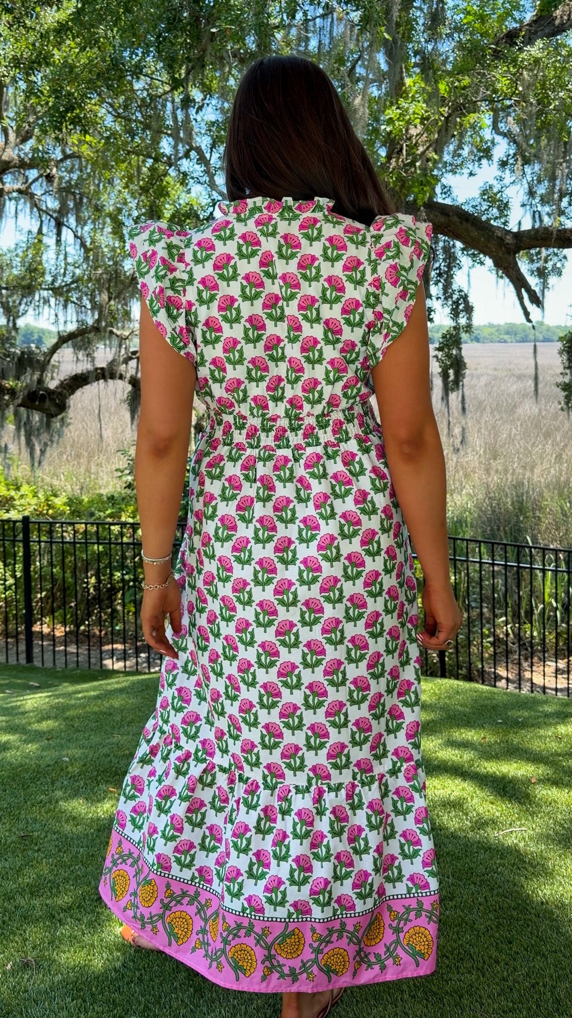 Woman wearing a floral dress standing in a garden with trees and greenery.