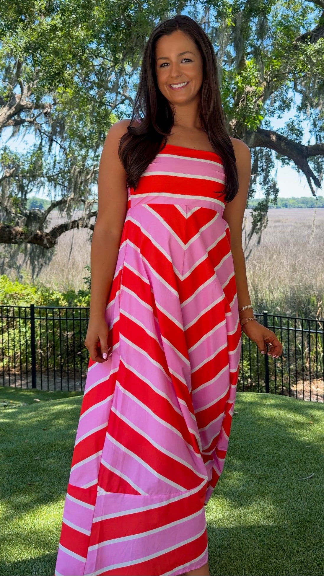 Woman wearing a red and pink striped dress standing outdoors with trees and grass in the background