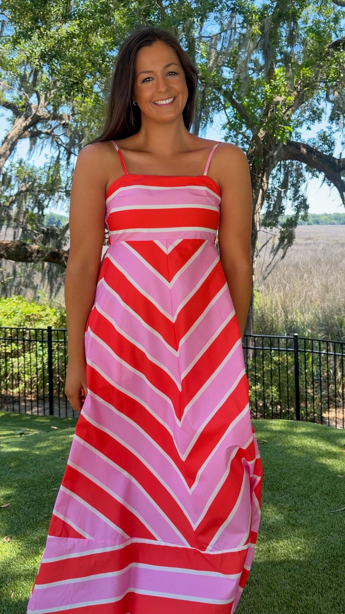 Woman wearing a red and pink striped dress standing outdoors with trees and grass in the background