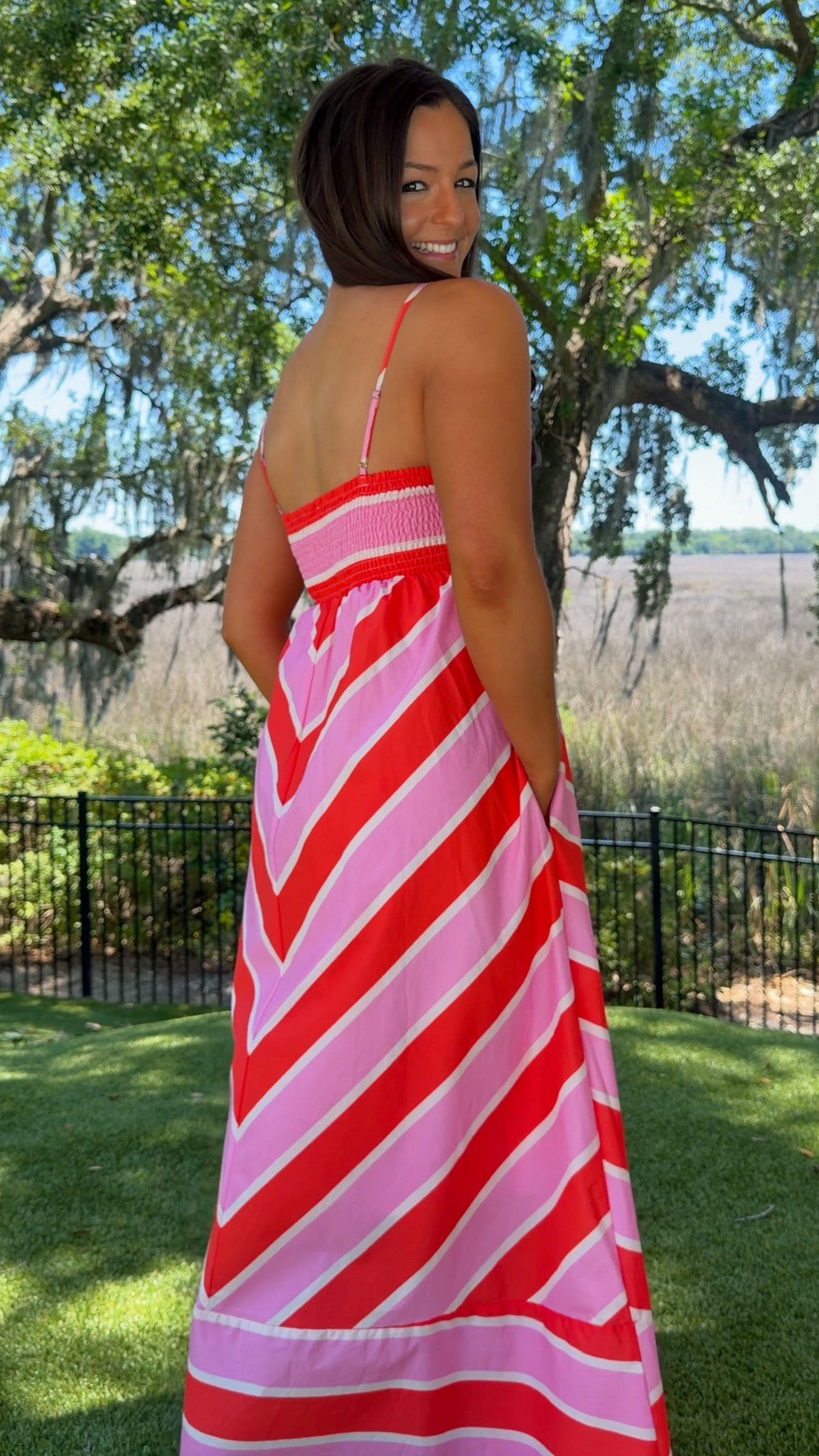 Woman wearing a red and pink striped dress standing outdoors with trees and grass in the background