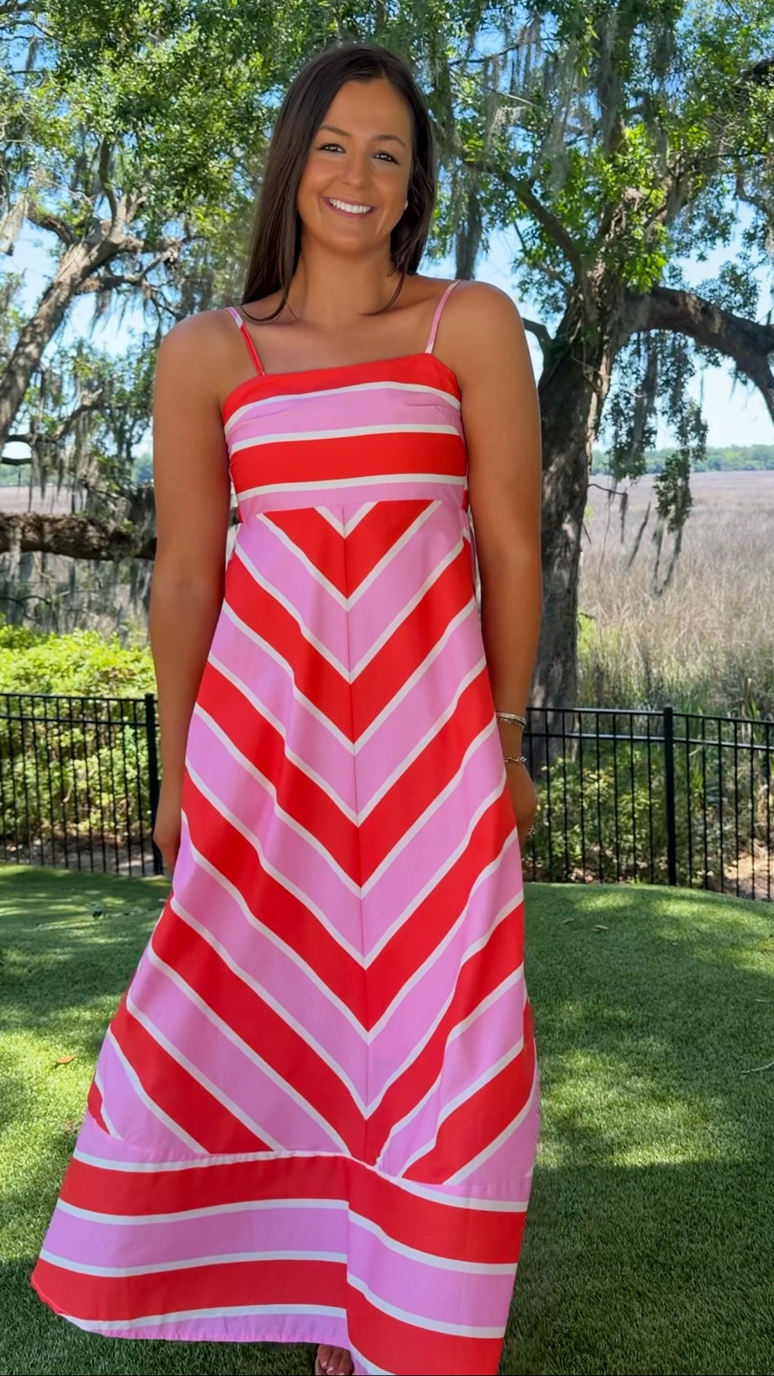 Woman wearing a red and pink striped dress standing outdoors with trees and grass in the background