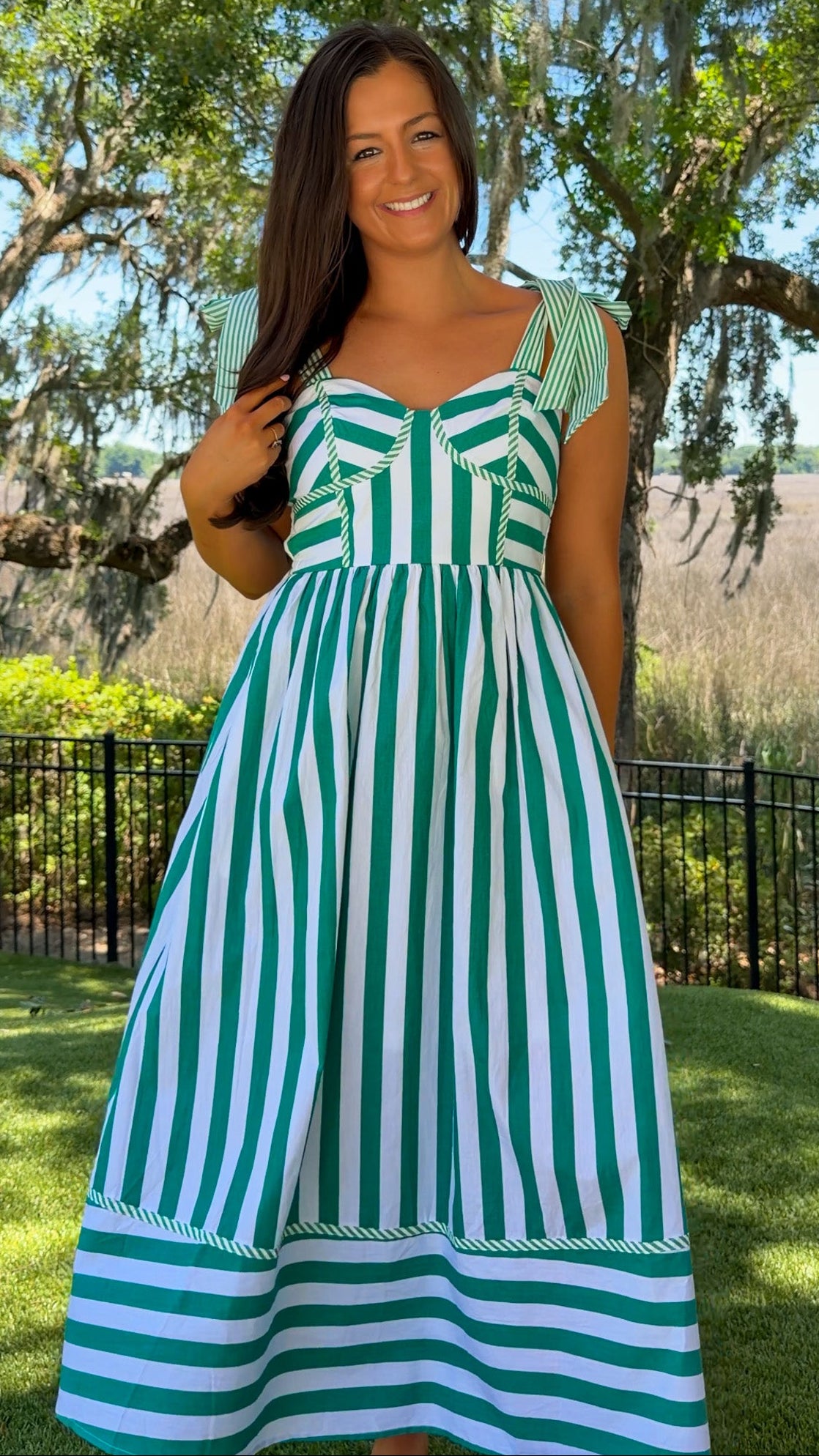 Woman wearing a green and white striped dress standing outdoors with trees and grass in the background
