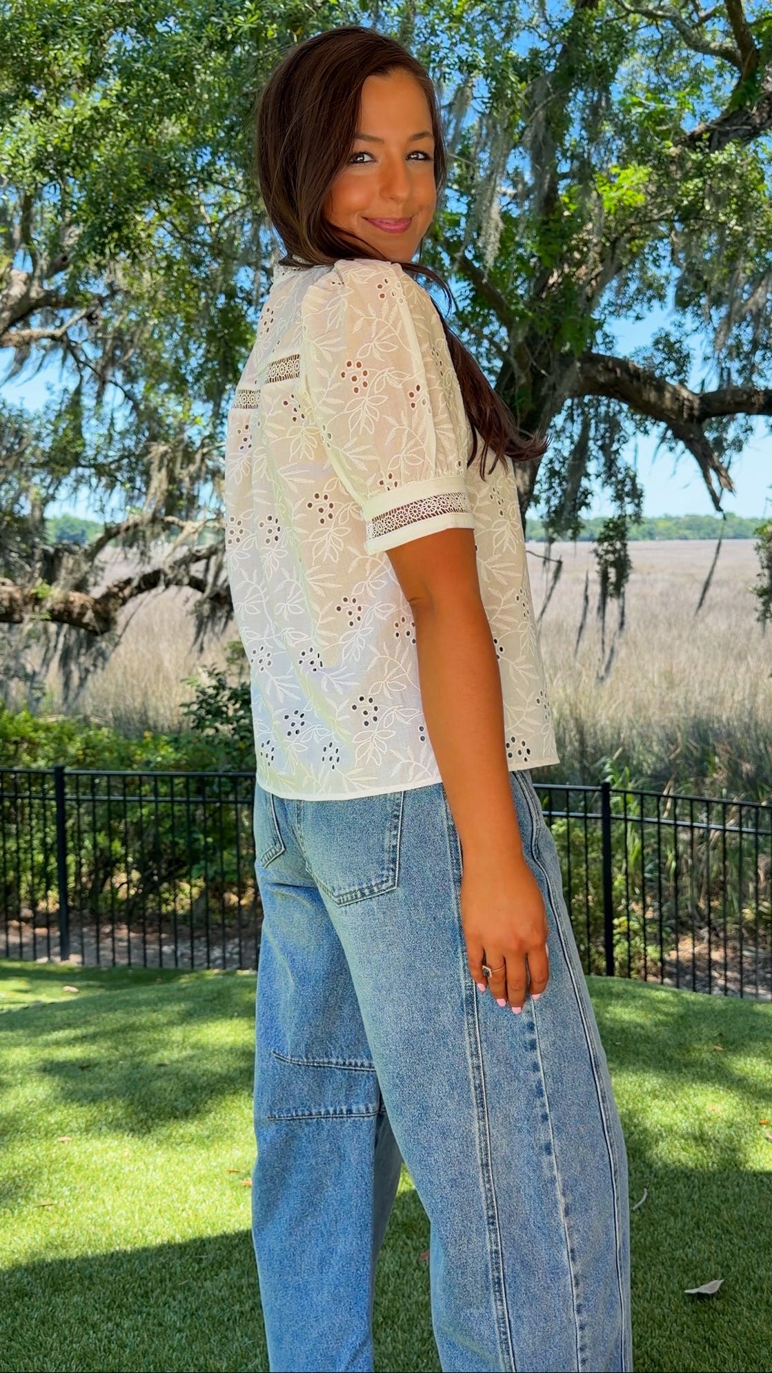 Woman wearing a white lace top and blue jeans standing outdoors with trees and grass in the background