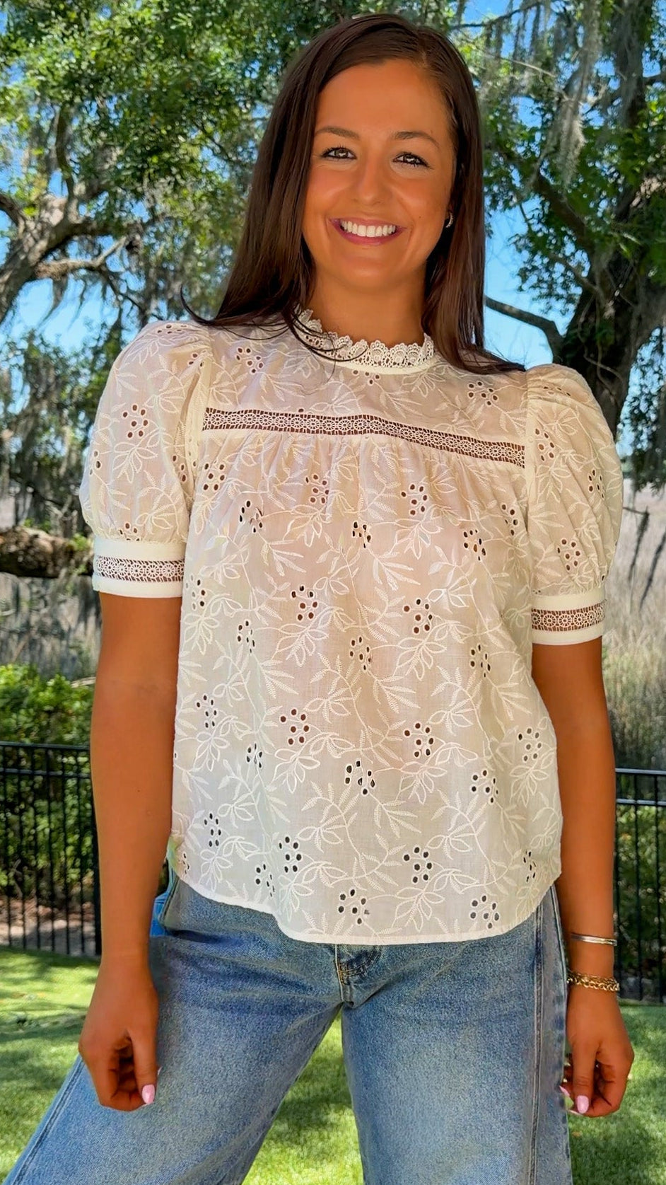 Woman wearing a white lace top and blue jeans standing outdoors with trees and grass in the background